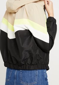 Chaqueta a bloques de color en beige, blanco, negro y amarillo neón. Cuenta con capucha, puños elásticos y un dobladillo fruncido. Material suave y ligero.