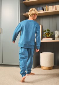 Plavi komplet od pamučne trenirke i hlača, s opuštenim krojem, okruglim izrezom i rebrastim manžetama. Glatka tkanina s minimalnim detaljima.