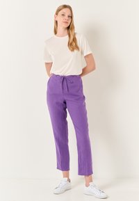 Paarse linnen broek met een elastische tailleband, gecombineerd met een eenvoudig wit t-shirt en witte sneakers. De stof heeft een textuurafwerking.