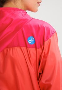 Veste imperméable rose avec une texture brillante, arborant un logo circulaire bleu sur l'épaule gauche. Design léger et sportif.