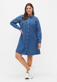 Jeanshemdkleid in Blau, mit durchgehender Knopfleiste, zwei Brusttaschen, langen Ärmeln und einem lockeren Schnitt. Weiche Textur und knielang.