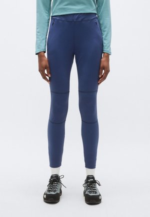 Personne portant un pantalon bleu marine ajusté avec des poches zippées, des chaussettes blanches, des chaussures de randonnée noires et grises, et un haut bleu clair à manches longues, debout.