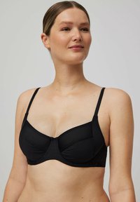 Mujer con piel clara y cabello recogido, llevando un sujetador negro con aros y detalles de panel de malla, mirando ligeramente hacia un lado.