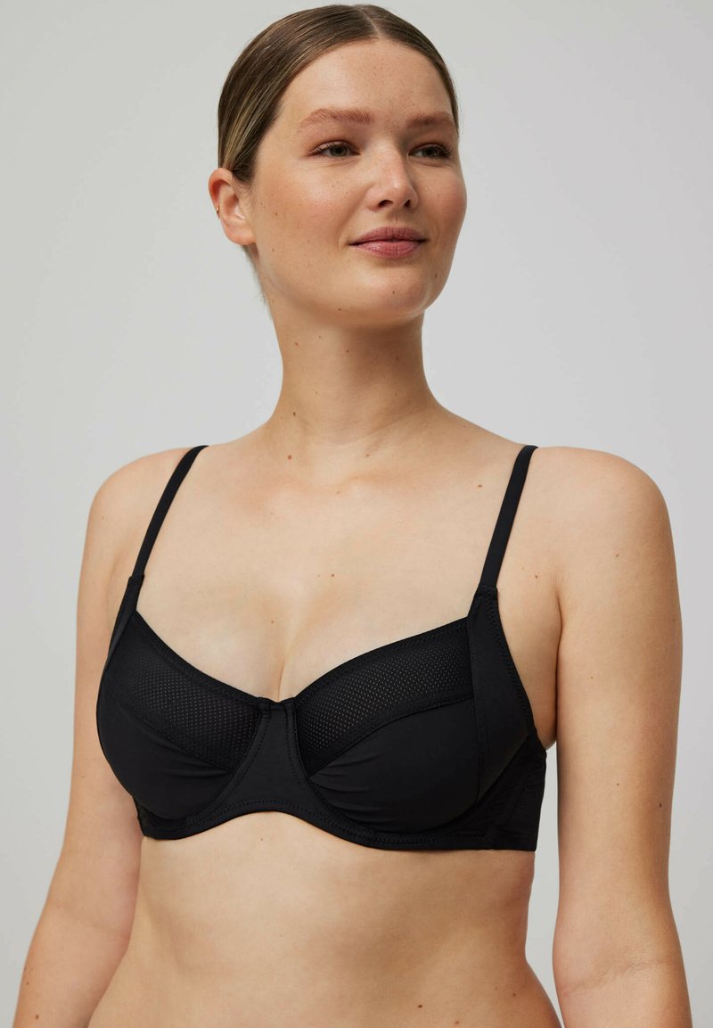 Mujer con piel clara y cabello recogido, llevando un sujetador negro con aros y detalles de panel de malla, mirando ligeramente hacia un lado.