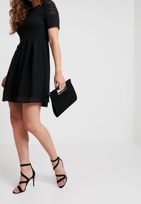 Schwarz lace Kleid mit kurzen Ärmeln, ausgestelltem Rock; kombiniert mit einer schwarzen Wildleder-Clutch und sandalen mit hohen Riemen. Glatte Textur, klassisches Design.