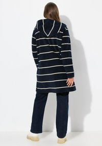 Felpa con cappuccio a righe blu navy con coulisse, caratterizzata da linee orizzontali bianche, tessuto testurizzato e spacchi ai lati dell'orlo. Indossata con pantaloni scuri.