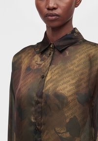 Gros plan d'une personne portant une chemise transparente boutonnée avec un col, présentant un motif abstrait marron et noir et un texte répété "Just Cavalli".
