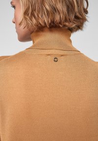 Pletací svetr s vysokým límcem v měkké zlatohnědé přízi s žebrovanou texturou, s knoflíkovým doplňkem na zadní straně.