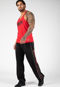 Red tank top with black logo text, paired with black sweatpants featuring red side text. Smooth fabric, athletic fit, and black sneakers.