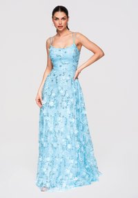 Femme portant une robe bleu clair sans manches, longue jusqu’au sol, avec des motifs floraux, posant avec une main sur la hanche sur un fond uni.