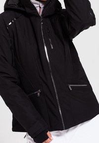 Svart isolerad jacka med dragkedjestängning, avtagbar huva och dragkedjefickor på sidorna. Har ribbad textur och stiliga silverdetaljer.