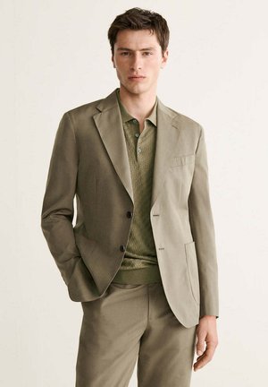 Joven con un traje beige y una camiseta polo verde, de pie con una mano en el bolsillo sobre un fondo claro y liso.