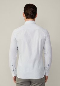 Biely košeľový dres s vertikálnymi modrými pruhmi, s gombíkovými manžetami a špicatým golierom. Zadný pohľad zobrazuje prispôsobený strih a hladkú štruktúru látky.