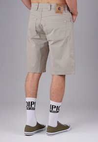 Beige cotton shorts with a straight cut, featuring a leather patch and copper rivets, paired with white logo socks and olive green sneakers.