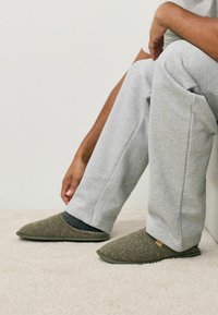 Grønne stofslipper med en tekstureret finish og mønstrede overlay, båret med grå sweatpants, hvilende på en lyst farvet overflade.