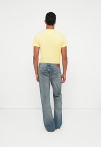 Lühikeste tumepruunide juustega mees, kes kannab heledat kollast T-särki ja blekitud siniseid teksapükse, seisab valge taustaga seljaga poole.