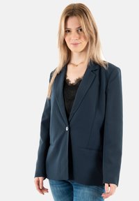 Blazer entallado azul marino con diseño estructurado, cierre de un botón, bolsillos de solapa y textura suave. Lleva puesto encima de una blusa de encaje negra.