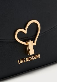 Μαύρη τσάντα χειρός από συνθετικό δέρμα με χρυσό κούμπωμα σε σχήμα καρδιάς και λογότυπο "LOVE MOSCHINO" με χρυσά γράμματα στο κάτω μέρος.