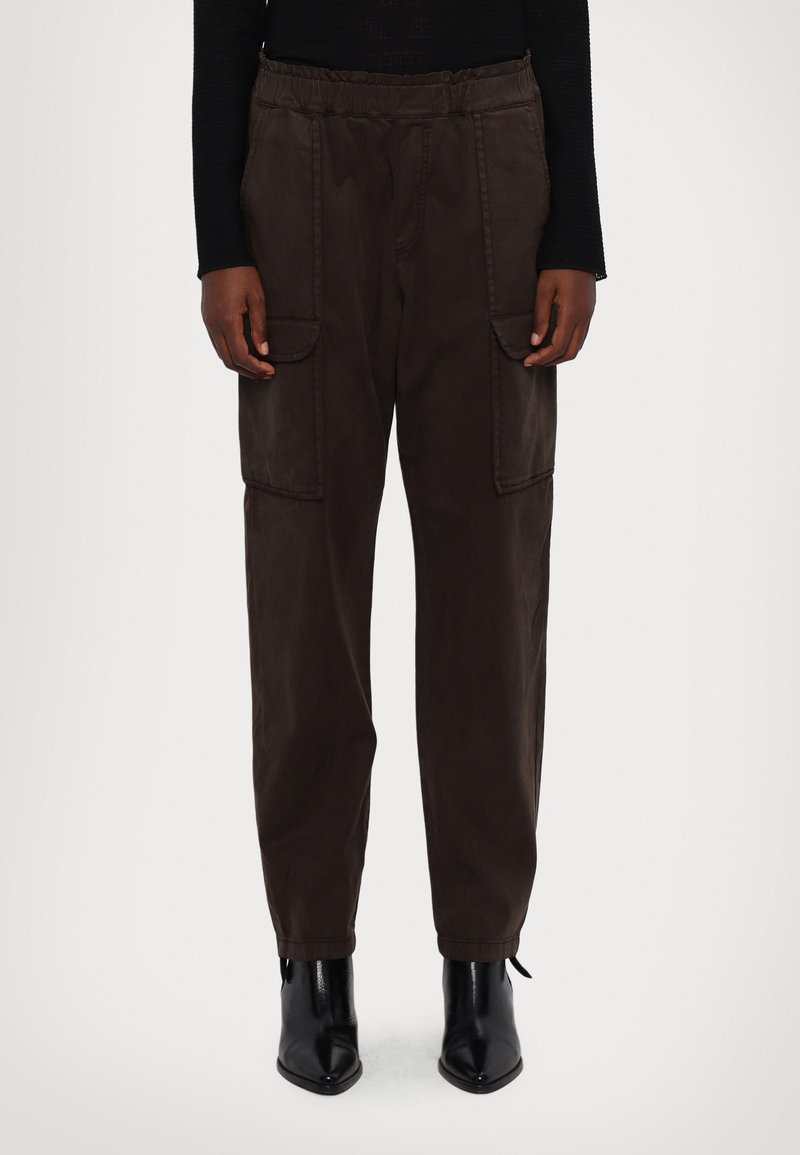 Personne portant un pantalon cargo marron foncé avec des poches à rabat devant et des bottines noires à bout pointu, debout devant un fond uni.