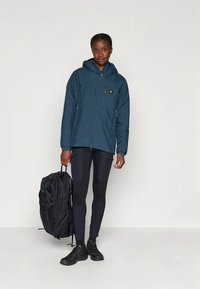 Tamsiai mėlyna, atspari vandeniui striukė su kapišonu, su užtraukiamais kišenėmis, derinama su juodais tamprėmis, juodu kupriniu ir juodais bateliais.