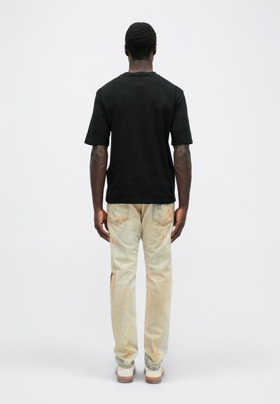 Tricou negru din bumbac cu mâneci scurte, asociat cu blugi bej decolorati, având pete portocalii și buzunare pe spate. Spatele modelului este vizibil.