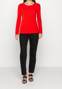 Rød langærmet bluse med rund halsudskæring, parret med sorte slim-fit bukser og sorte strop sandaler, sat mod en neutral baggrund.