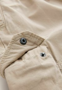 Tessuto beige con texture morbida, caratterizzato da due bottoni a pressione neri e cuciture decorative. Il primo piano mette in risalto il tessuto e i dettagli metallici.