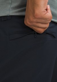 Tmavé námornícké nohavice s hladkou textúrou majú bočné vrecko a zapínanie na gombík. Ruka sa drží vrecka, čo ukazuje flexibilitu látky.