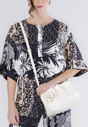 Žena oblečená v černo-bílo-béžovém topu a kalhotách s vzorem, držící bílou kabelku s květinovým detailem a ramenním popruhem.