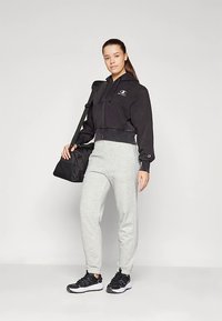 Jauna moteris juodu užtrauktuku užsegamu džemperiu, pilkomis kelnėmis ir juodomis sportinėmis batelėmis, ant peties nešanti juodą sportinę rankinę prieš paprastą foną.