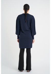 Abito lavorato a maglia blu navy con maniche a sbuffo, lunghezza al ginocchio, abbinato a pantaloni neri a coste svasati. Design minimalista, colori solidi, texture liscia.