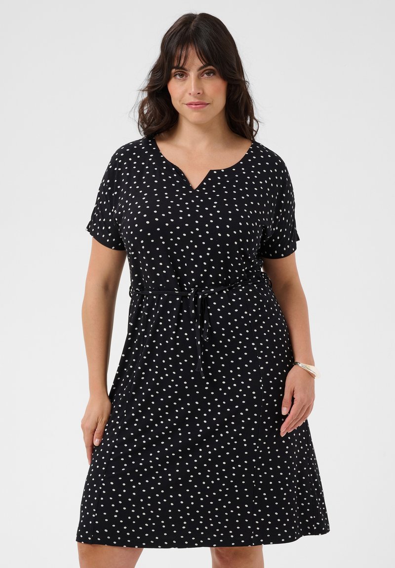 Femme portant une robe noire à manches courtes avec des pois blancs et une ceinture à la taille, debout devant un fond clair uni.
