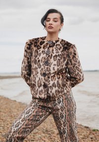 Leopárdmintás műszőrme kabát fekete gombokkal és magas gallérral, hozzáillő, testhezálló kialakítású, ugyancsak leopárdmintás nadrággal párosítva.