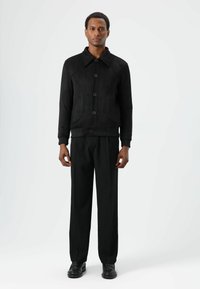 Zwarte suède jas met een kraag, knoopsluiting aan de voorkant en geribde manchetten. Gecombineerd met loszittende zwarte broek en zwarte schoenen voor een monochrome uitstraling.