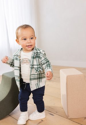 Peuters in een groen-witte geruite hoodie over een licht shirt, marineblauwe broek en witte schoenen, staande nabij zachte, abstracte zitmeubels.