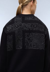 Černá mikina s texturovaným vzorem na zadní straně, která má abstraktní motivy v světle šedé barvě. Vyrobená z měkkého materiálu s žebrovanými manžetami.