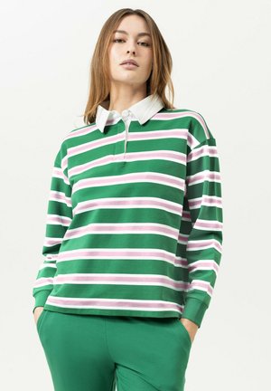 Vrouw met steil schouderlang haar die een groen rugbyshirt met lange mouwen draagt met witte en roze strepen en een groene broek, handen in de zakken.