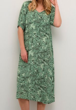 Femme portant une robe verte longueur genoux avec un motif feuillu noir et des manches au coude, debout contre un mur clair uni.