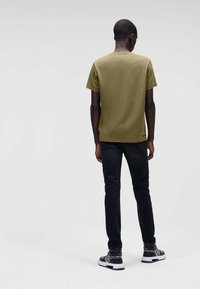 Olivgrünes T-Shirt mit kurzen Ärmeln, einfarbiger Stoff, lässige Passform, kombiniert mit schwarzen Slim-Fit-Jeans und gemusterten Sneakers. Rückansicht.
