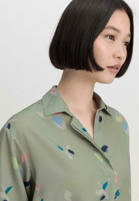 Helle grüne Bluse mit Kragen und einem mehrfarbigen abstrakten Muster. Glatte Textur mit lockerer Passform.