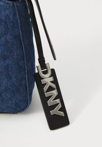 Τσάντα από τζιν με σκούρο μπλε υφή, με μαύρη δερμάτινη ετικέτα που έχει ασημένια γράμματα "DKNY", συνδεδεμένη με μια ταιριαστή μαύρη λουρίδα.