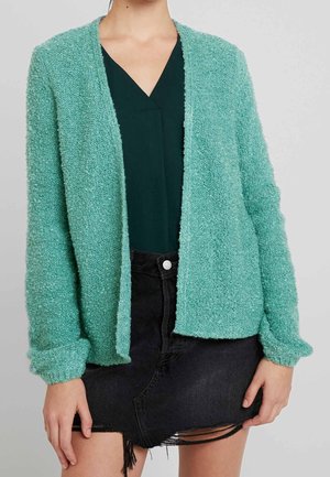 Mujer lleva un cárdigan abierto texturizado color verde azulado sobre una blusa de cuello en V verde oscuro y una minifalda de mezclilla negra desgastada.