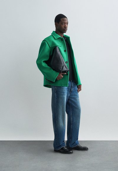 Chaqueta de mezcla de lana verde con botones negros, vaqueros de pierna ancha azules y un bolso de mano de cuero negro, con un fondo neutro.