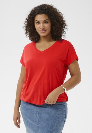 Femme aux cheveux bouclés portant un T-shirt rouge à col en V et une jupe en jean bleue, debout devant un fond blanc uni.