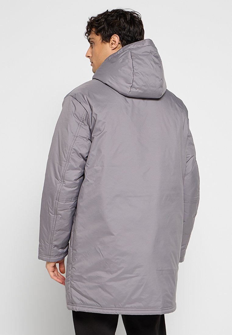 Hombre vestido con un abrigo acolchado gris de longitud media con capucha, de pie de espaldas a la cámara frente a un fondo sencillo.