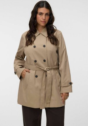 Femme aux longs cheveux foncés porte un trench-coat beige à double boutonnage noué à la taille avec des poches, pantalon noir, debout devant un fond uni.