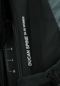 Schwarzer Rucksack mit einem auffälligen weißen Textlabel "DUCAN SPINE 28-35 FRAUEN." Enthält Maßangaben für Größenanpassungen.
