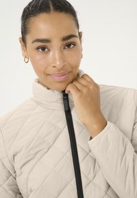 Veste matelassée beige clair avec un col haut et une fermeture éclair noire. Présente une surface texturée et des manches légèrement bouffantes.