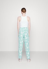 Kvinna som står vänd bort, iförd ett vitt ärmlöst linne, ljusblå byxor med vit palmträdsmönster och ljusa slip-on skor.