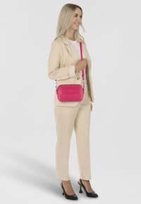 Borsa a tracolla sintetica rosa con logo in rilievo, ferrature argentate e tracolla regolabile, abbinata a un completo beige e tacchi neri.